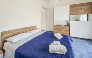 Ein Schlafzimmer mit einem Bett, einem Holz-Kopfteil, einer blauen Bettdecke und einem weißen Kopfkissen. Ein Holzschrank und ein Spiegel befinden sich an der Wand.