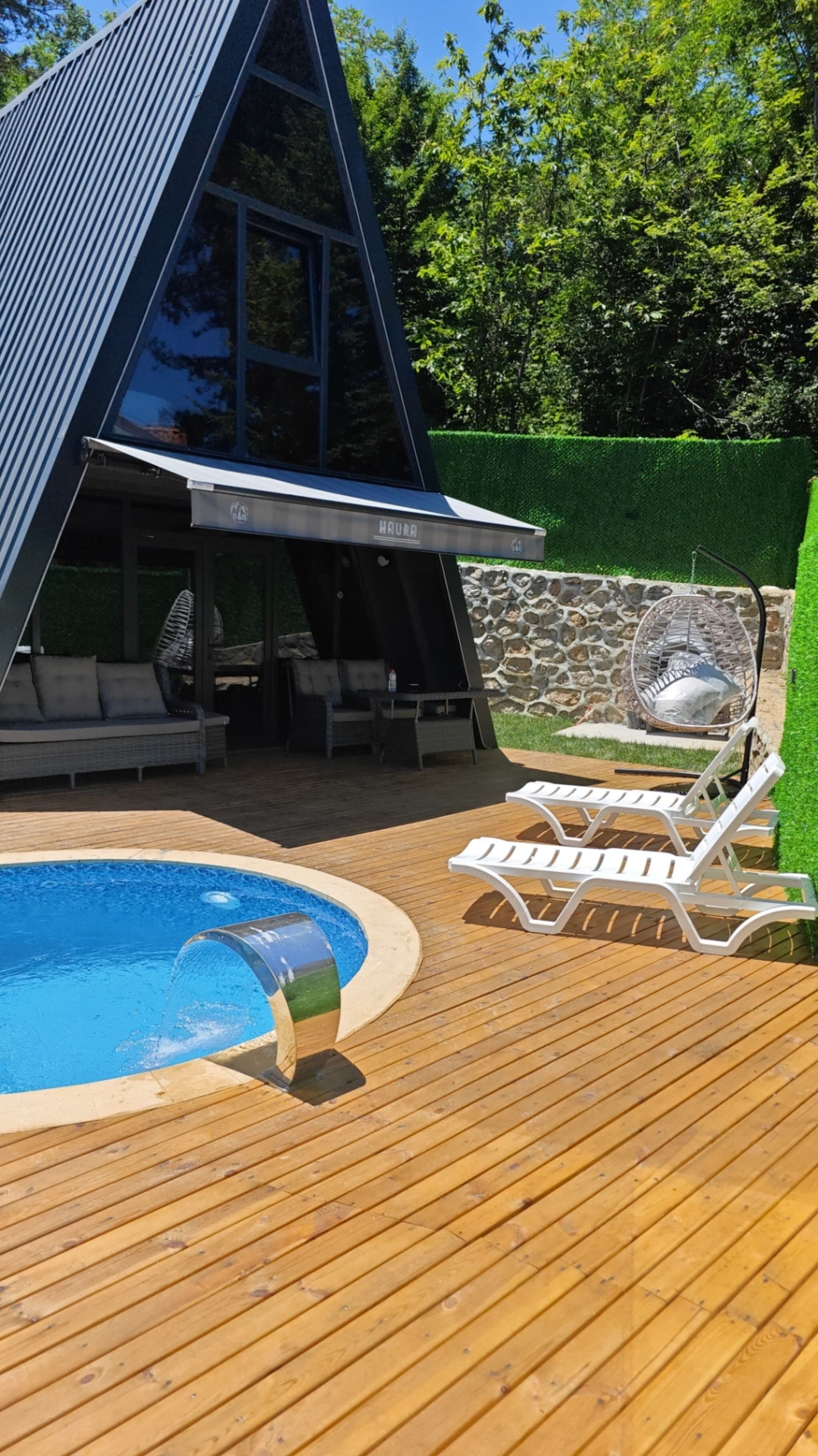 A modern outdoor pool area with a wooden deck, lounge chairs, and a stone wall.