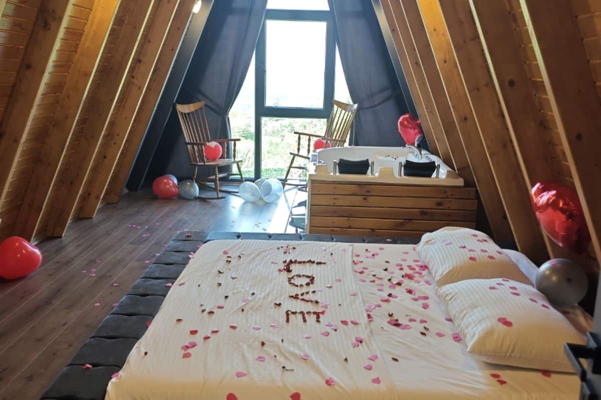 A cozy attic bedroom with a bed, wooden beams, and a view through a large window.