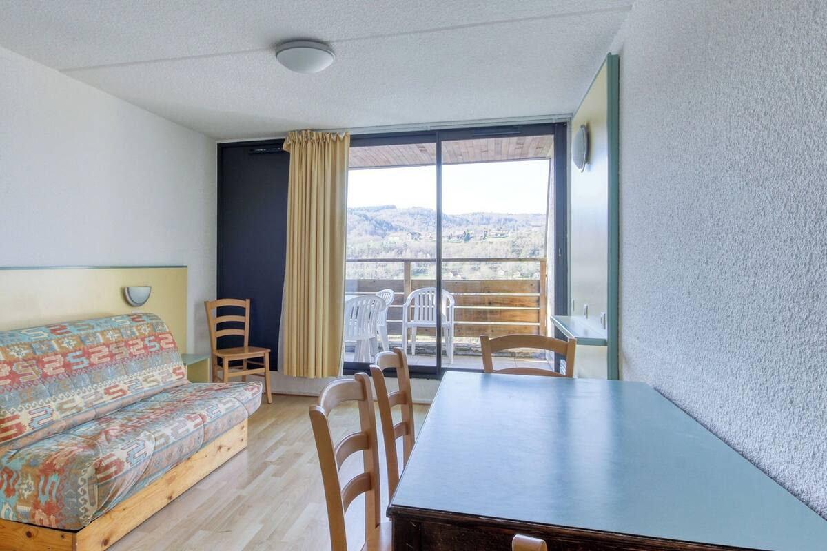 A room with a sofa, a dining table, chairs, a window with a view of the outdoors, and a ceiling light.