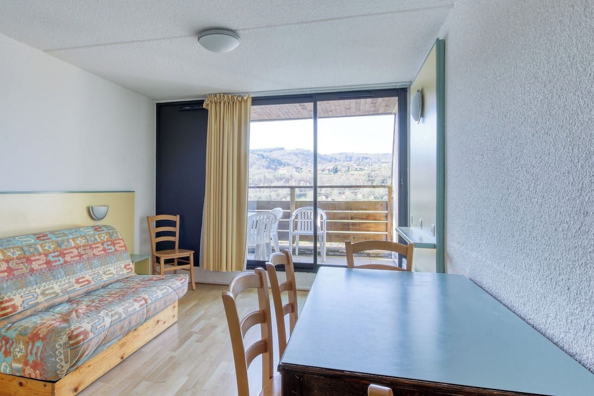 Una habitación con sofá, una mesa de comedor, sillas, una ventana con vistas al exterior y una lámpara en el techo.