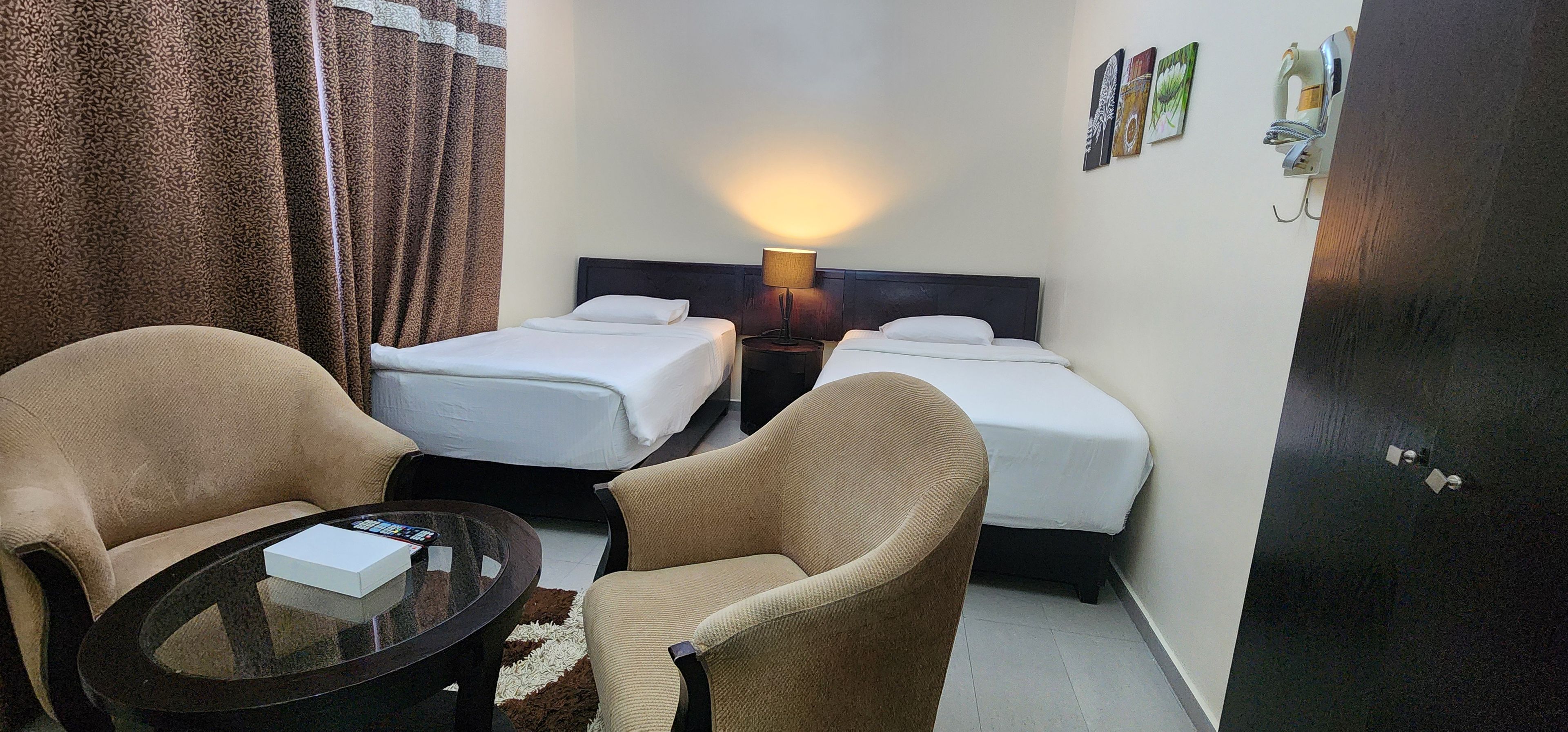 Quarto de hotel com duas camas de solteiro, uma mesa redonda com um controle remoto, duas poltronas, um abajur e uma porta com fechadura.