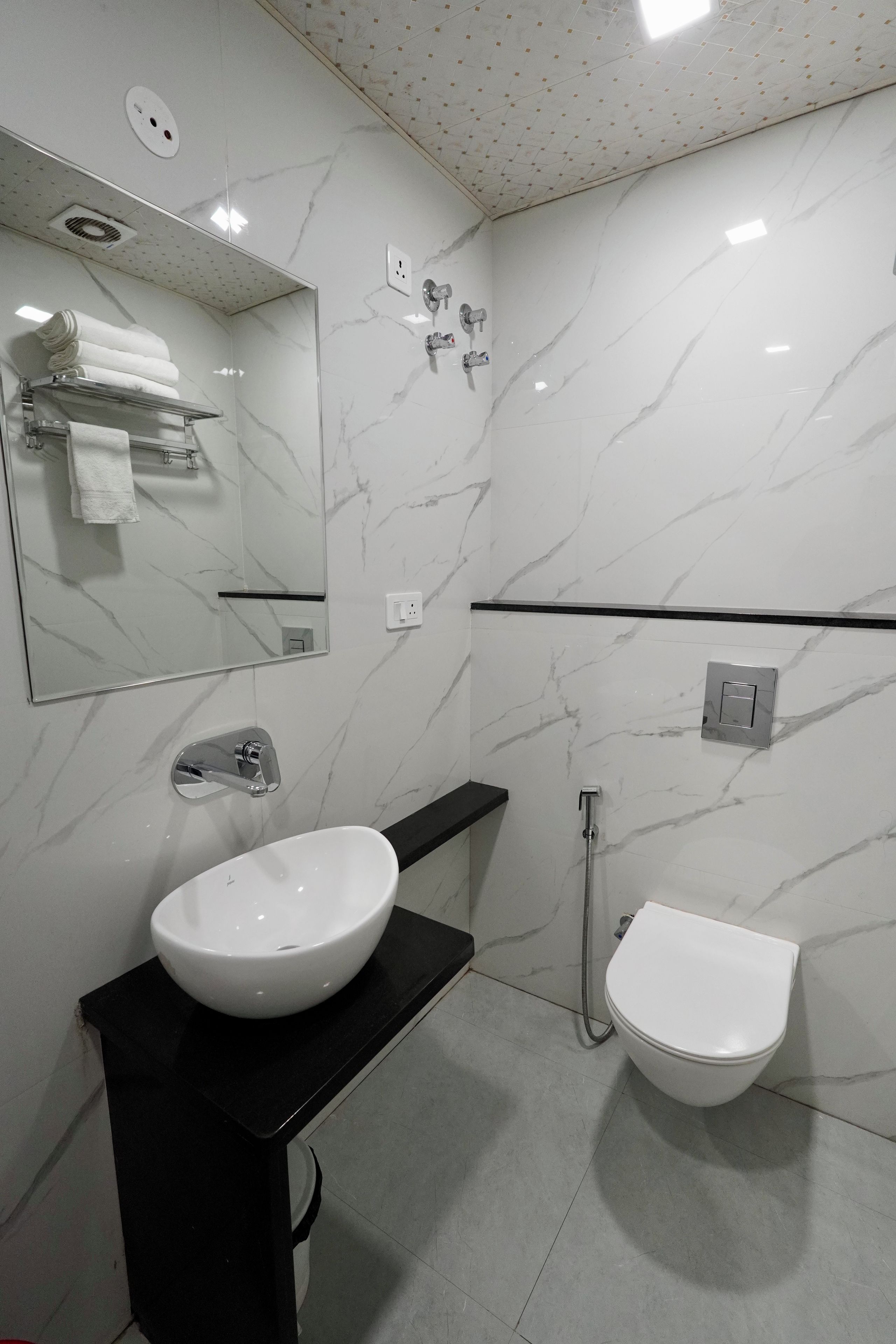 A modern bathroom with a white marble wall, a black countertop, a white sink, a toilet, a mirror, and a shower.