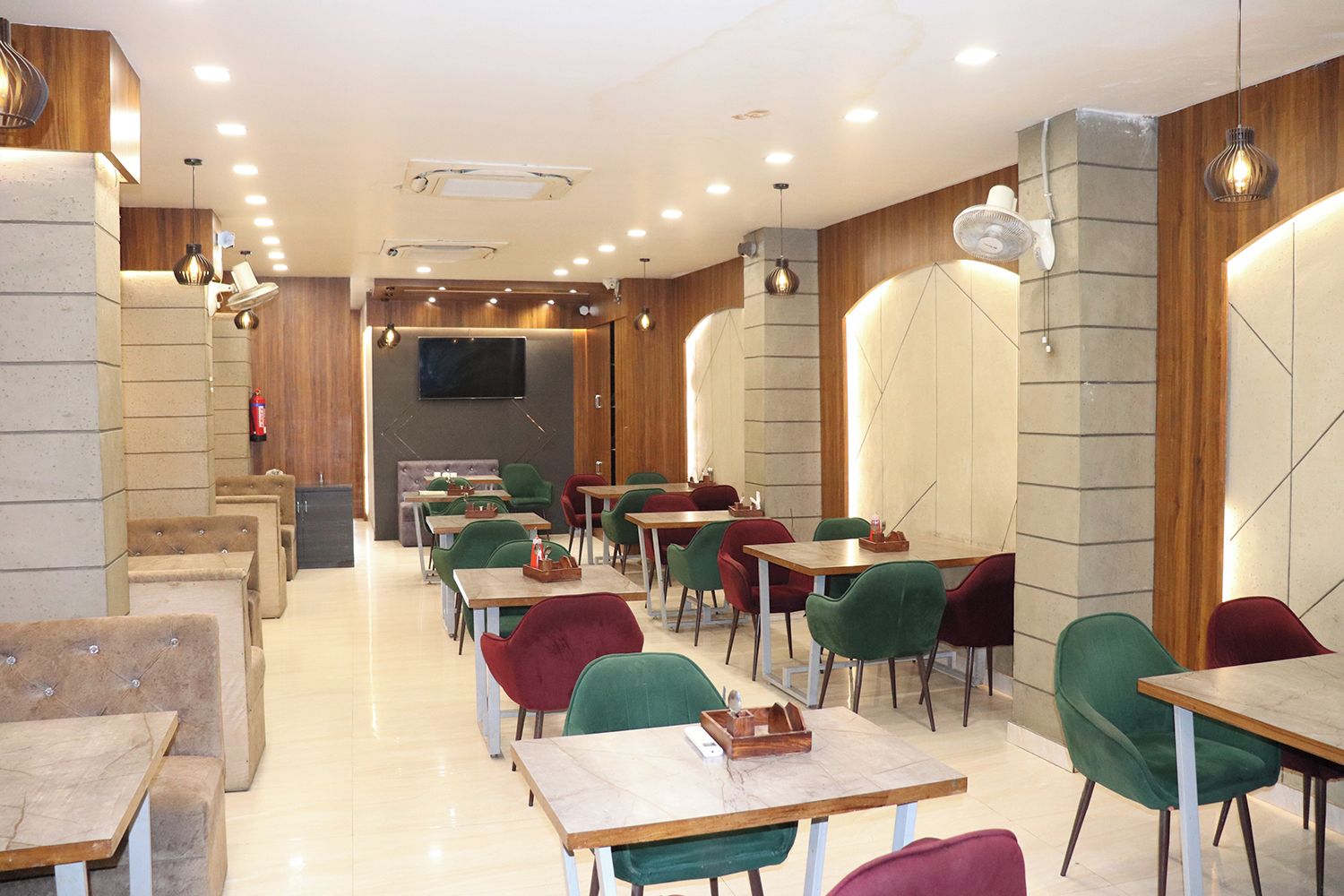 A modern restaurant interior with multiple tables and chairs, a flat-screen TV mounted on the wall, and recessed ceiling lighting.