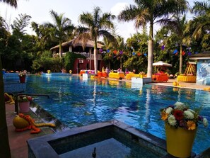 En swimmingpool med en byggnad i bakgrunden som har ett tak av halmtak, omgiven av palmer och färgstarka dekorationer.