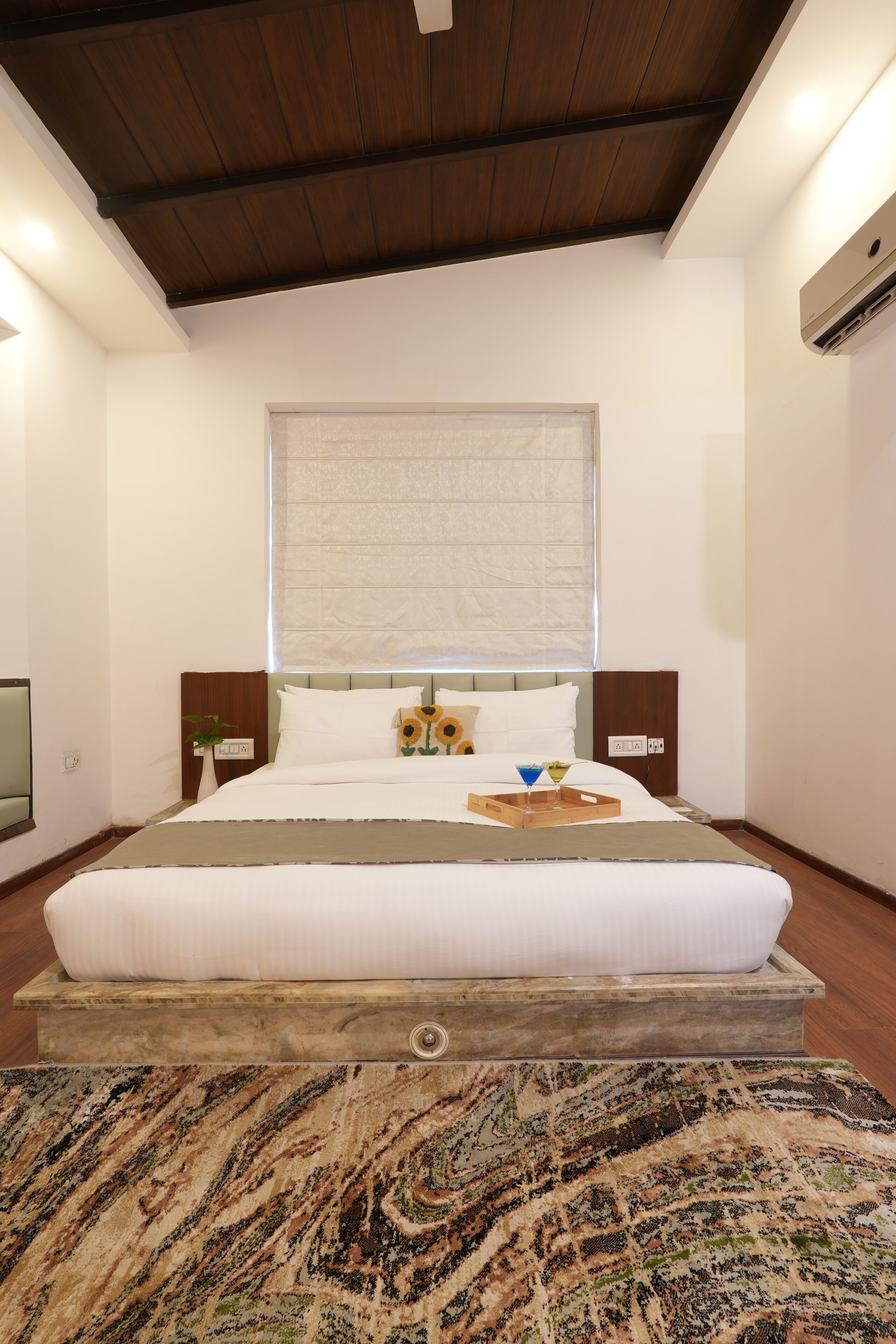 A bedroom with a large bed, a wooden headboard, a window with blinds, a bedside table with a lamp, and a wall-mounted air conditioner.