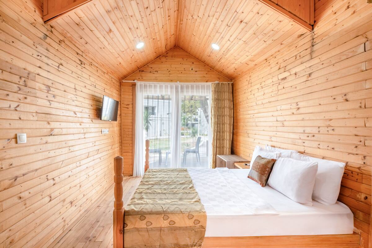 A wooden cabin room with a bed, a small table, and a TV mounted on the wall.