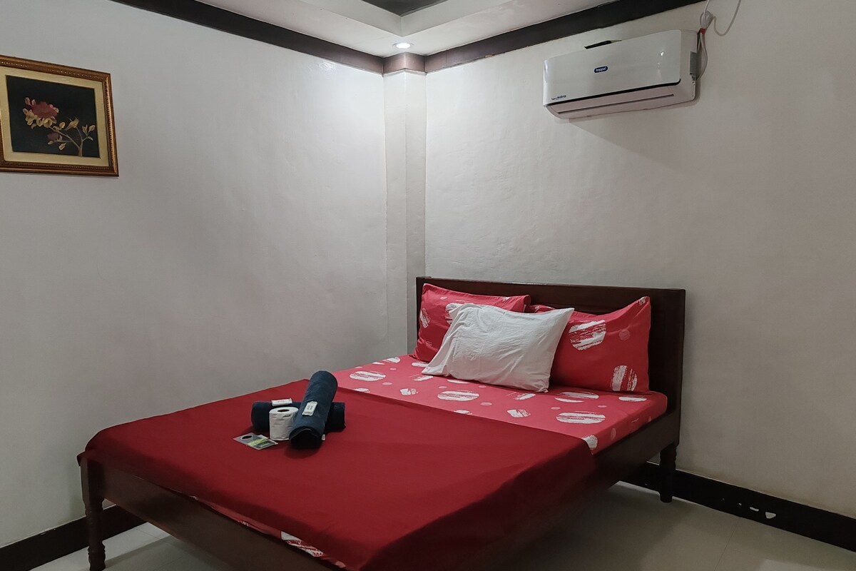 Una cama individual con una colcha roja, un almohadón blanco y un almohadón azul. Encima de la cama, hay un cuadro en la pared.