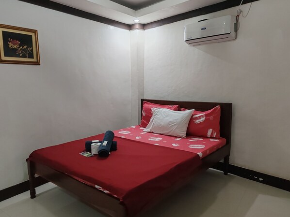 Una cama individual con una colcha roja, un almohadón blanco y un almohadón azul. Encima de la cama, hay un cuadro en la pared.