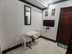 A room with a white table, two white chairs, a black-framed mirror, a mounted TV, and a wooden door. - Jace's Travellers Inn By Zuzu (El Nido)