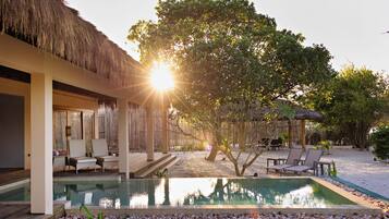 Un área junto a la piscina con sillas de descanso, una estructura de techo de paja y un árbol grande.