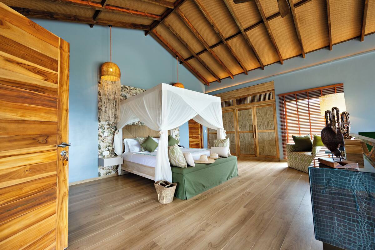 A room with a bed under a canopy, wooden flooring, a woven ceiling, and a view of the outside through a window.