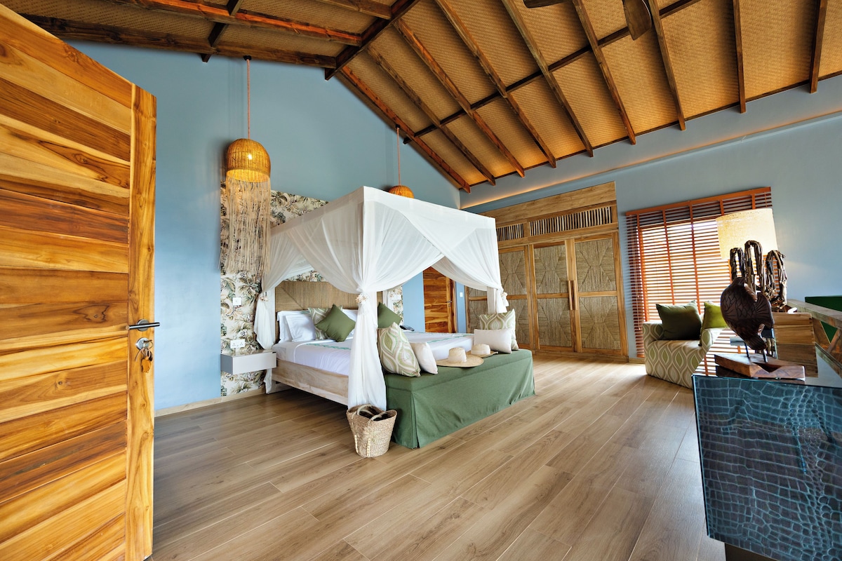 A room with a bed under a canopy, wooden flooring, a woven ceiling, and a view of the outside through a window.