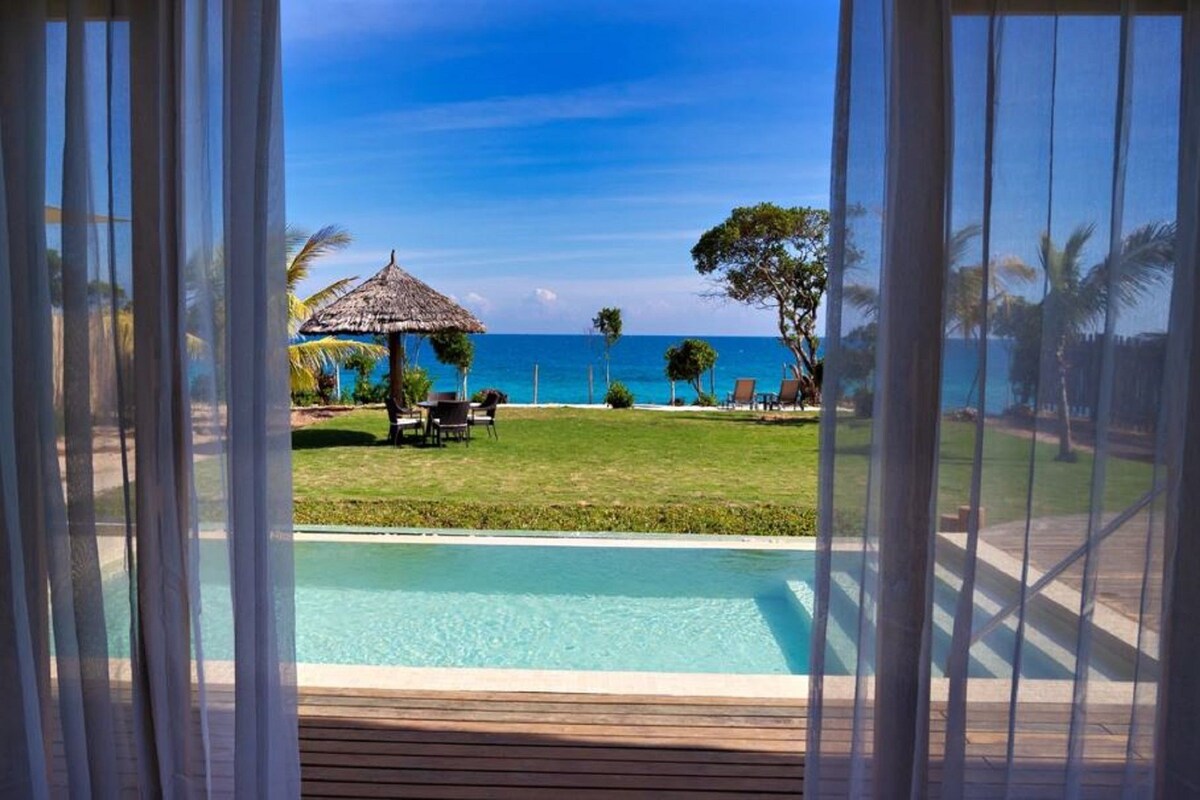 A pool with a view of the ocean, a thatched umbrella, and a grassy area with chairs.