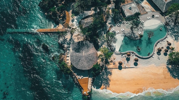 Vista aèria d'un complex turístic amb platja, piscina i una construcció amb teulada de palla.