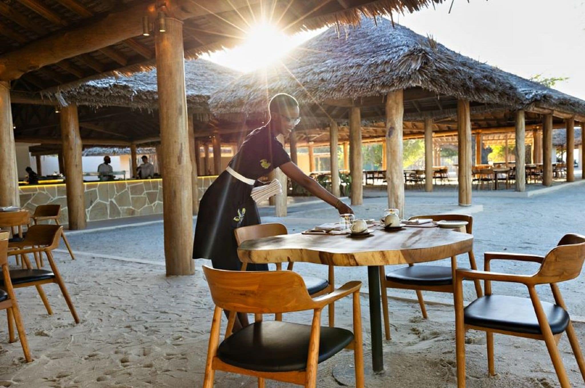 An outdoor dining area with wooden tables and chairs, thatched roof structures, and a sandy floor.
