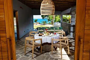 A dining area with a table set for four, surrounded by wicker chairs, under a woven ceiling light, with a view of the sea and greenery outside. - Bawe Island Resort (Bawe Island)