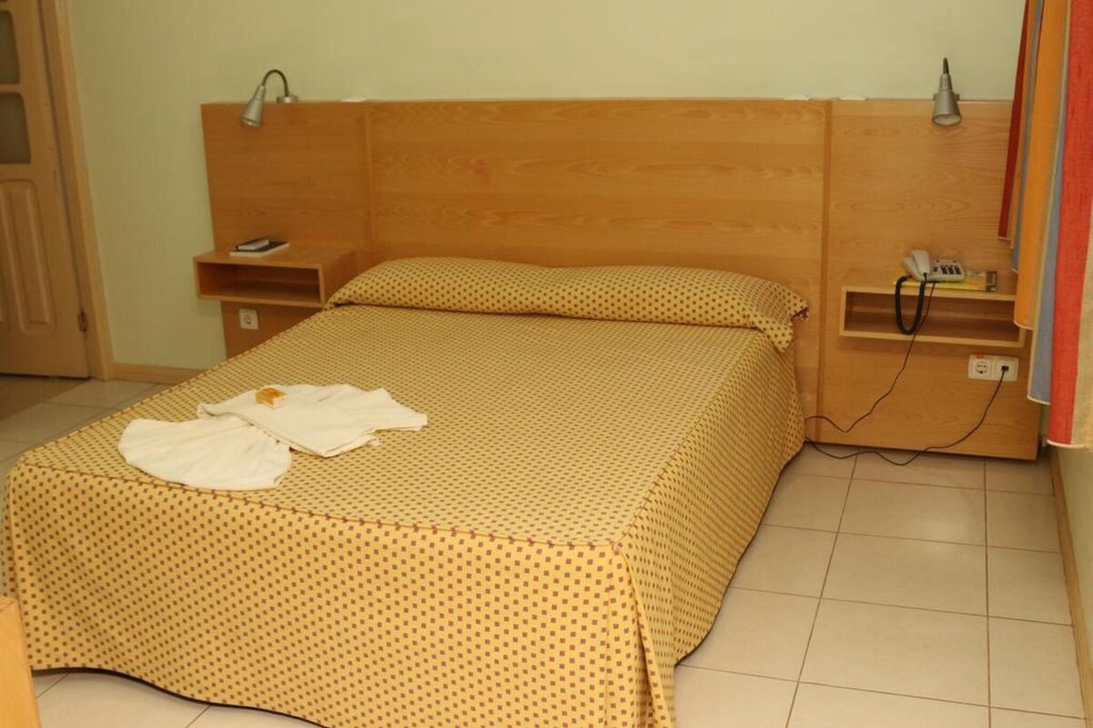 A single bed with a wooden headboard, a bedside table with a lamp, a phone, and a shelf with a lamp.