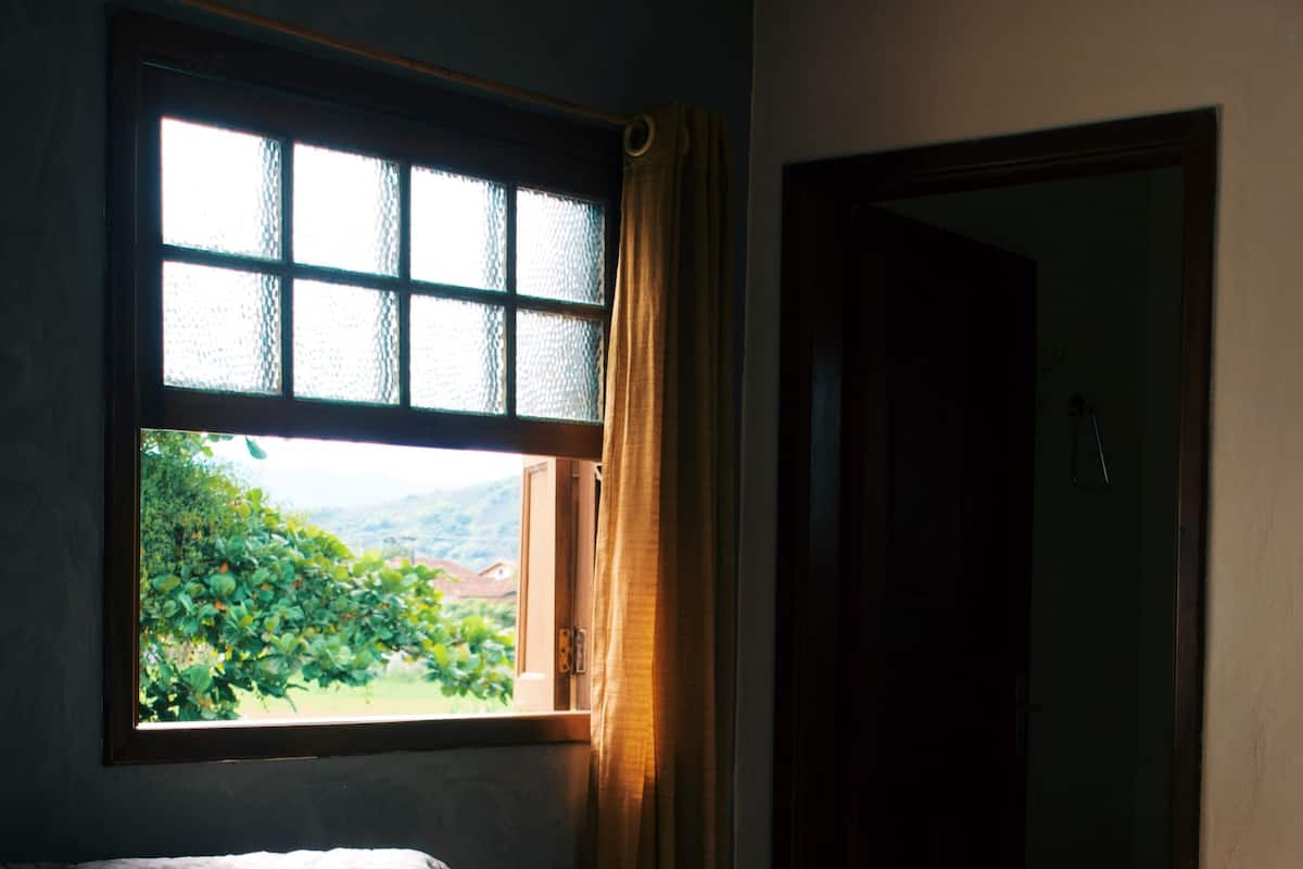 A room with a bed, a window with curtains, and a view of greenery outside.