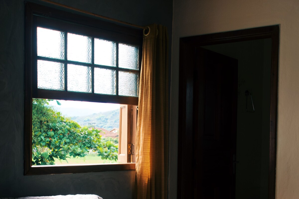 A room with a bed, a window with curtains, and a view of greenery outside.
