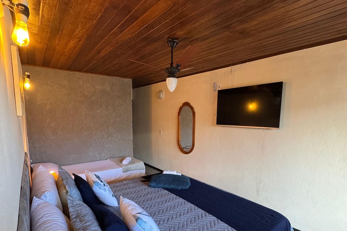 A bedroom with a bed, pillows, a ceiling fan, a wall-mounted TV, and a wooden ceiling.