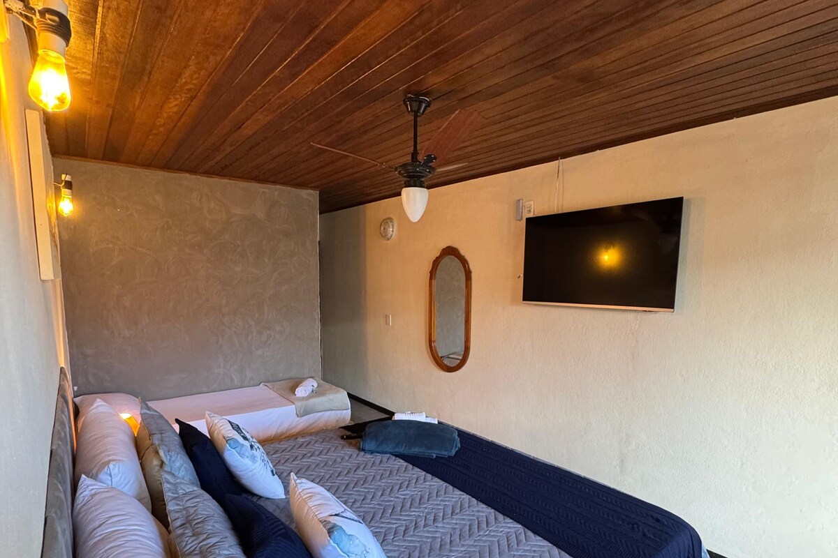 A bedroom with a bed, pillows, a ceiling fan, a wall-mounted TV, and a wooden ceiling.