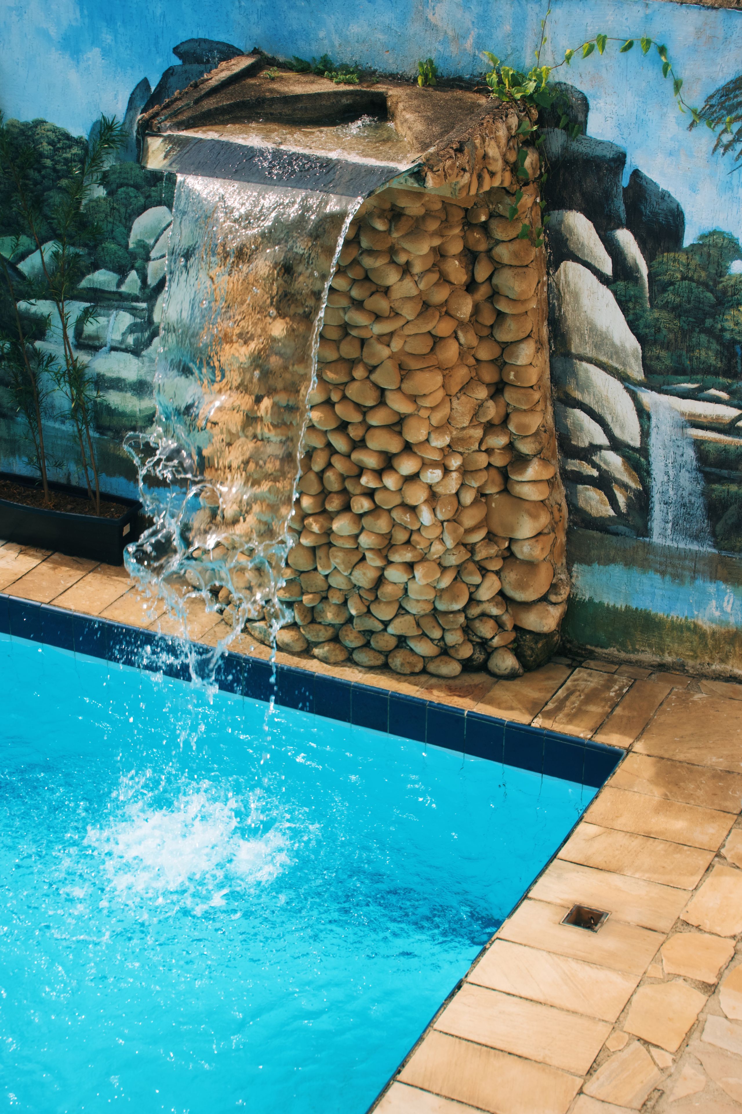 Ein Pool mit klarem, blauem Wasser, eine Steinmauer mit Wasserfall und ein Wandgemälde einer Naturlandschaft.