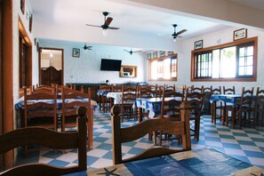 A dining area with wooden chairs and tables, a checkered floor, ceiling fans, and a television mounted on the wall. - Pouso De Icaro (Paraty)