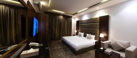 A modern hotel room with a large bed, a sofa, a TV, and a desk with a phone and a box.