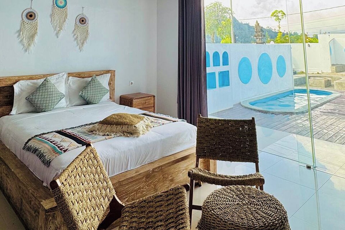 A bedroom with a bed, bedside tables, a chair, a view of a swimming pool, and a balcony.