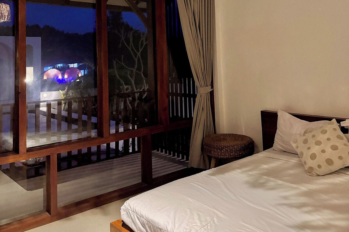 A bedroom with a large window, a bed with white bedding, a wooden headboard, and a small wicker stool.
