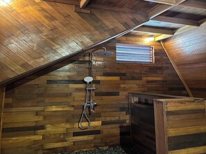 Ein Holzinnenraum mit schrägverdeckter Decke, einer Dusche mit Handbrause und einem Fenster mit Jalousien.
