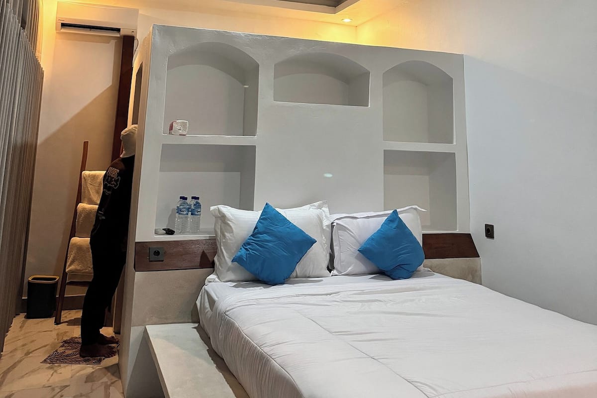 Quarto de hotel com uma cama, mesa de cabeceira, prateleira com garrafas e porta-toalhas.