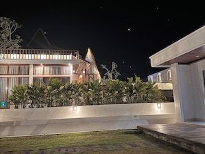 A modern building with a balcony, a well-lit entrance, and a garden area with plants.