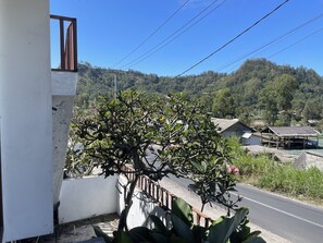 A balcony with a view of a road, a tree with pink flowers, and a hilly landscape in the background. - Volcano Side Kintamani Bali By Zuzu (Kintamani)