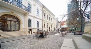 A cobblestone street with outdoor seating, a building with large windows, and a tree. - Jais Home-moon Apartment-muzeului Square-old Town (Cluj-Napoca)