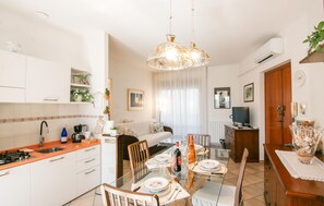 Een moderne keuken met witte kasten, een eettafel voor vier personen en een zithoek met een bank en televisie.