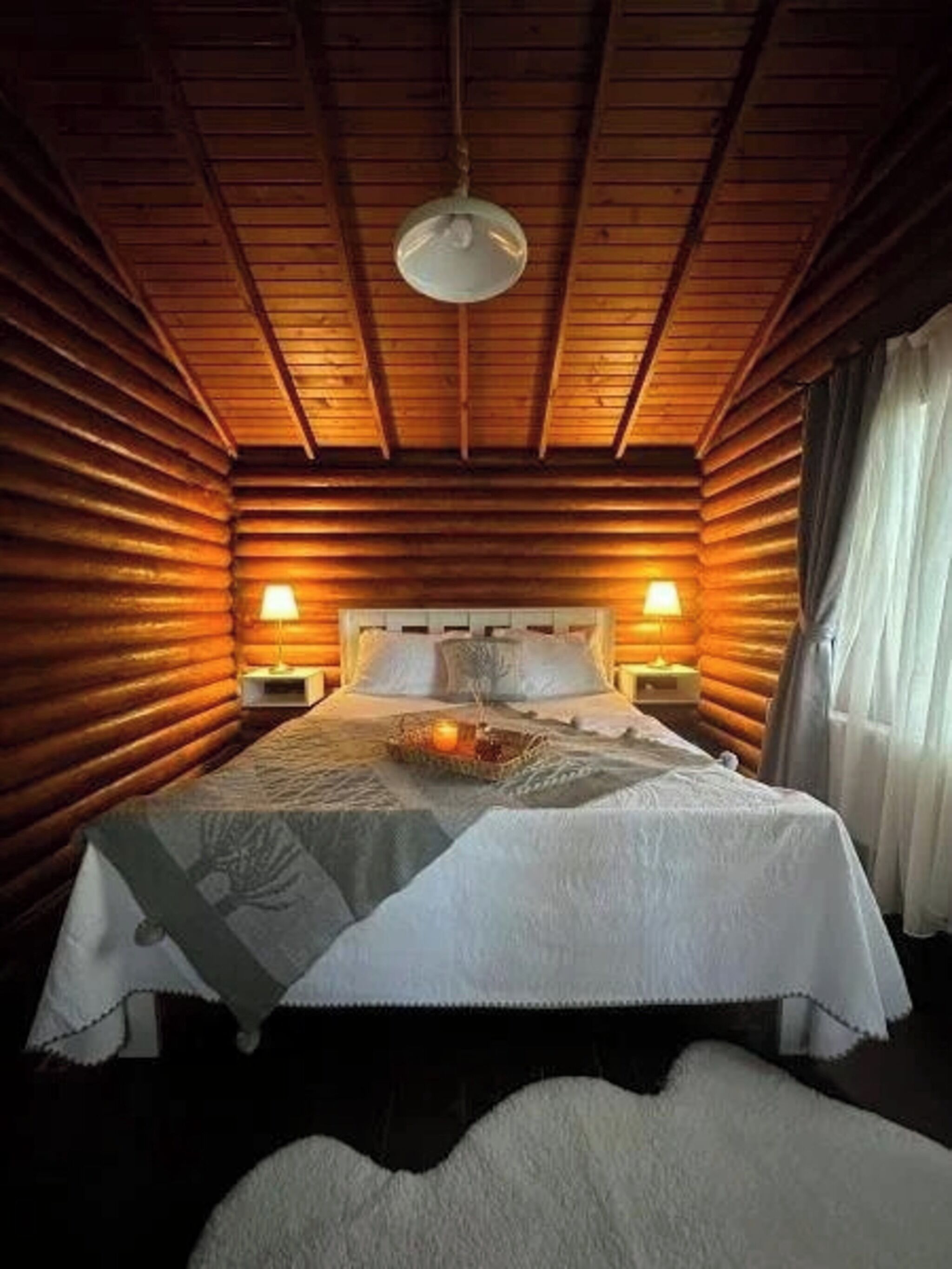 A log cabin-style bedroom with a bed, two bedside lamps, a hanging light fixture, and a window with curtains.