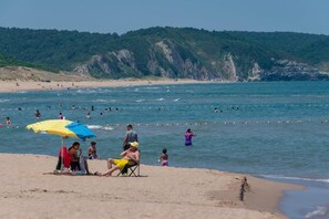 Ein Strand mit badenden und sonnenden Personen, ein klarer Himmel und ein bewaldeter Hügel im Hintergrund.