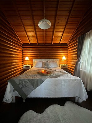 Ein Schlafzimmer in einer Blockhütte mit einem Bett, zwei Nachttischlampen, einer Pendelleuchte und einem Fenster mit Vorhängen.