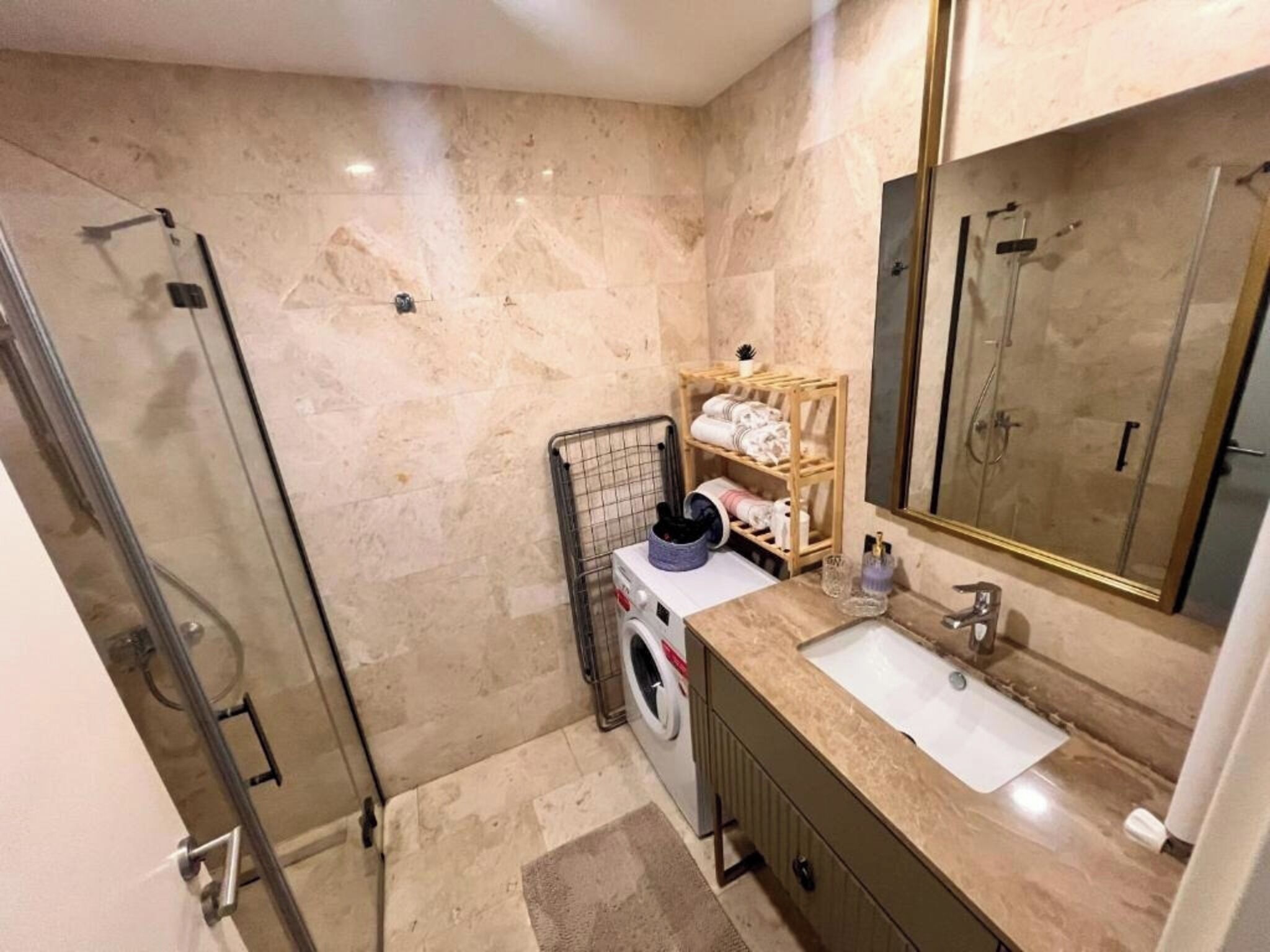 Een badkamer met een glazen douche, een wasmachine, een houten plank met handdoeken en een grote spiegel.