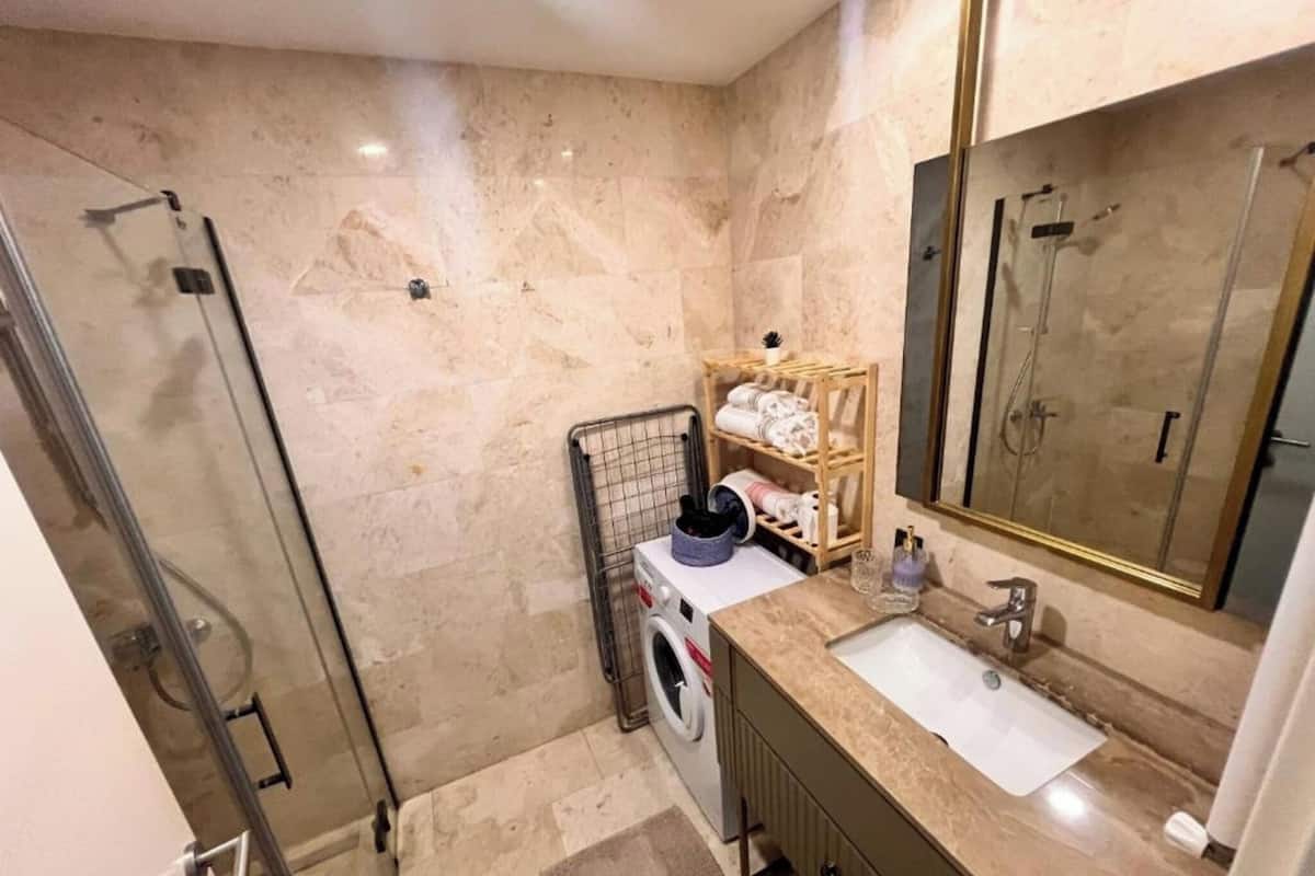 Ein Badezimmer mit Glasdusche, einer Waschmaschine, einem hölzernen Ablagefach mit Handtüchern und einem großen Spiegel.