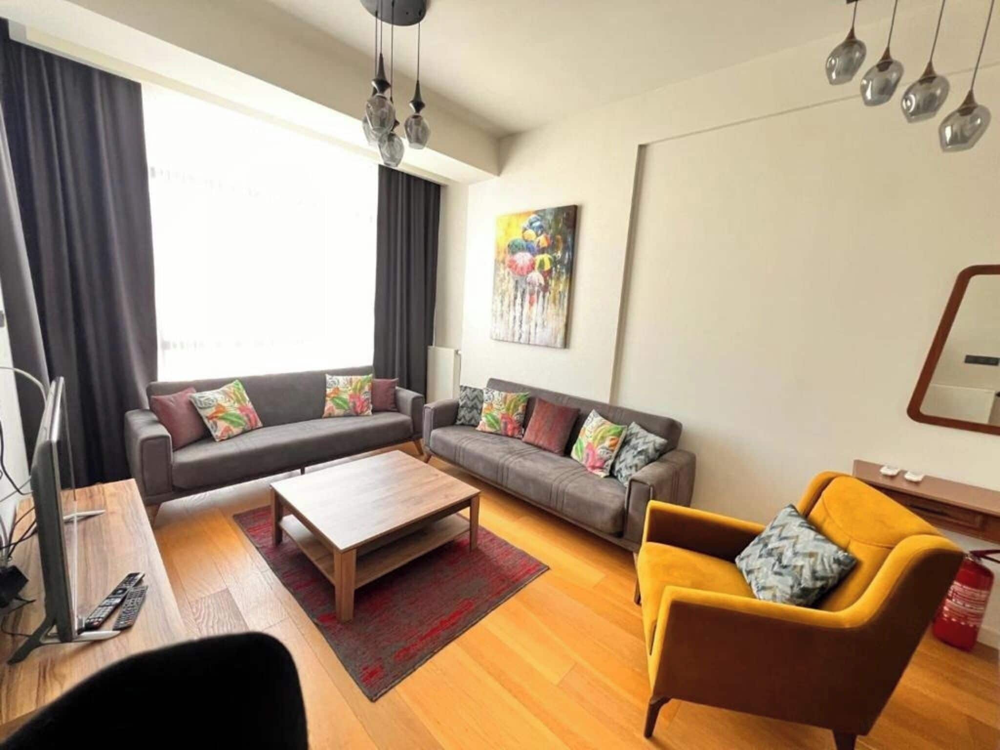 Een woonkamer met een zithoek, een salontafel, een gele fauteuil en een schilderij aan de muur.