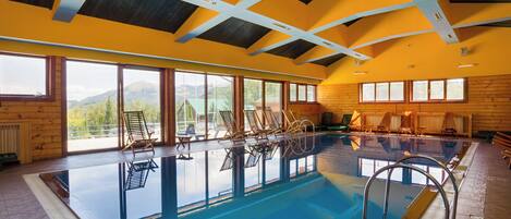 An indoor swimming pool with wooden deck chairs, large windows, and a high ceiling.
