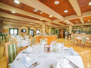 Un restaurante con techos de madera, varias mesas de comedor con manteles blancos y cristalería, y una zona de bar con estanterías llenas de botellas.