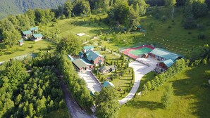 An aerial view of a rural property with multiple buildings, a sports court, and a winding road. - Hotel & Resort Gacka - Imanje Rakočević (Mojkovac)