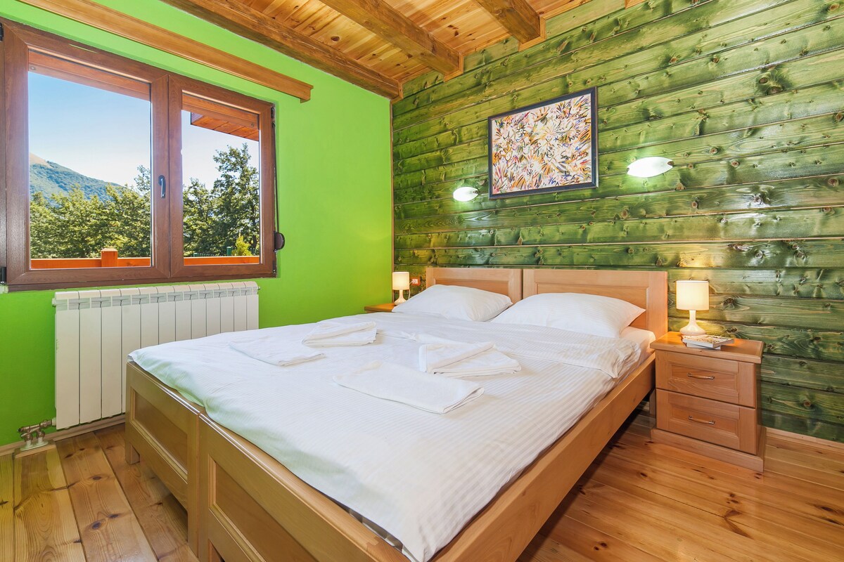 Ein hölzernes Bett mit weißen Bettwäsche und Handtüchern, ein Nachttisch mit Lampe sowie ein Fenster mit Blick auf Bäume und Berge.