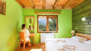 A bedroom with a wooden ceiling, a bed with white linens, a desk with a chair, a window with a view of trees, and a radiator. - Hotel & Resort Gacka - Imanje Rakočević (Mojkovac)