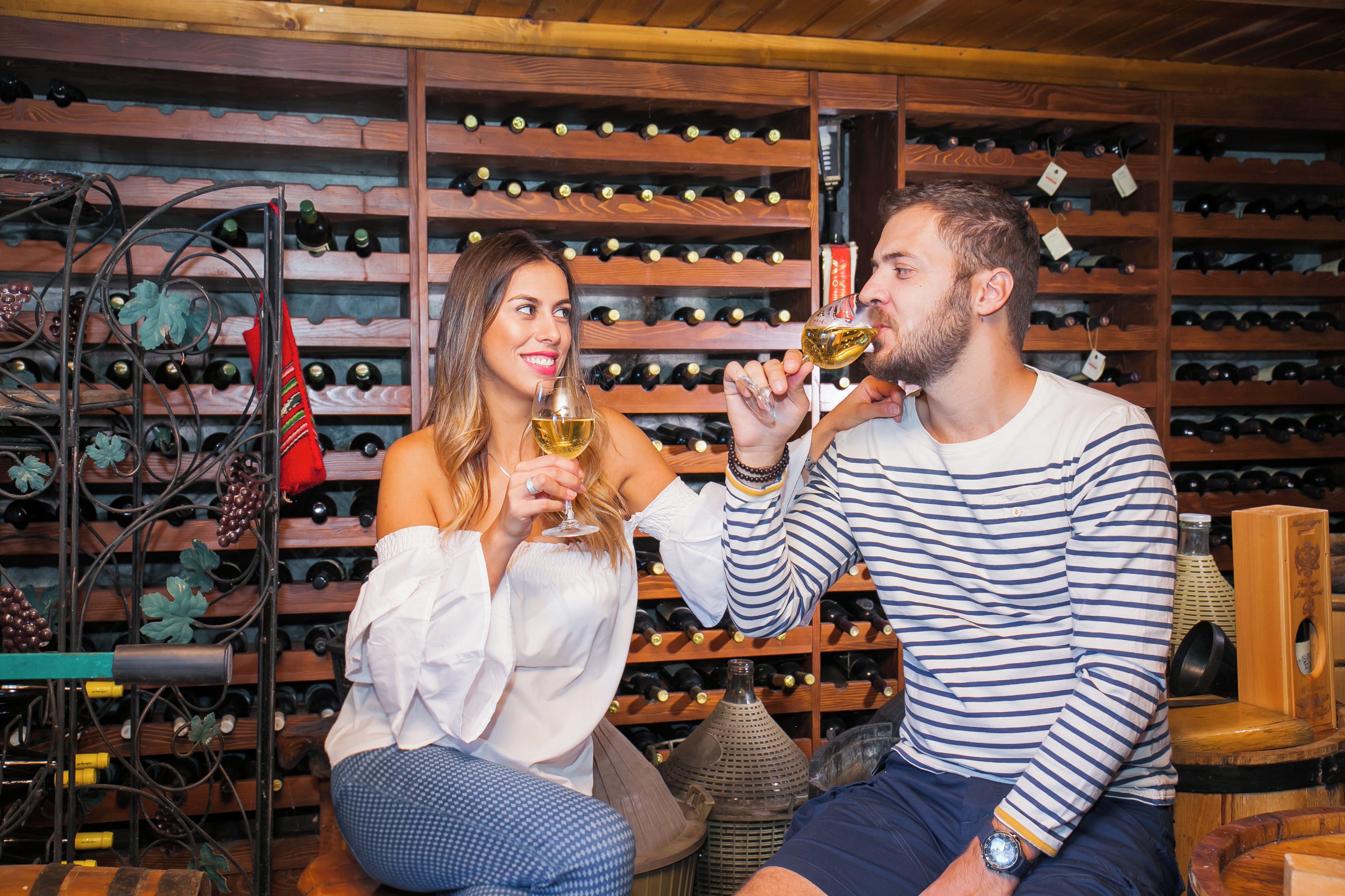 A man and a woman sitting in a wine cellar with wine racks in the background.