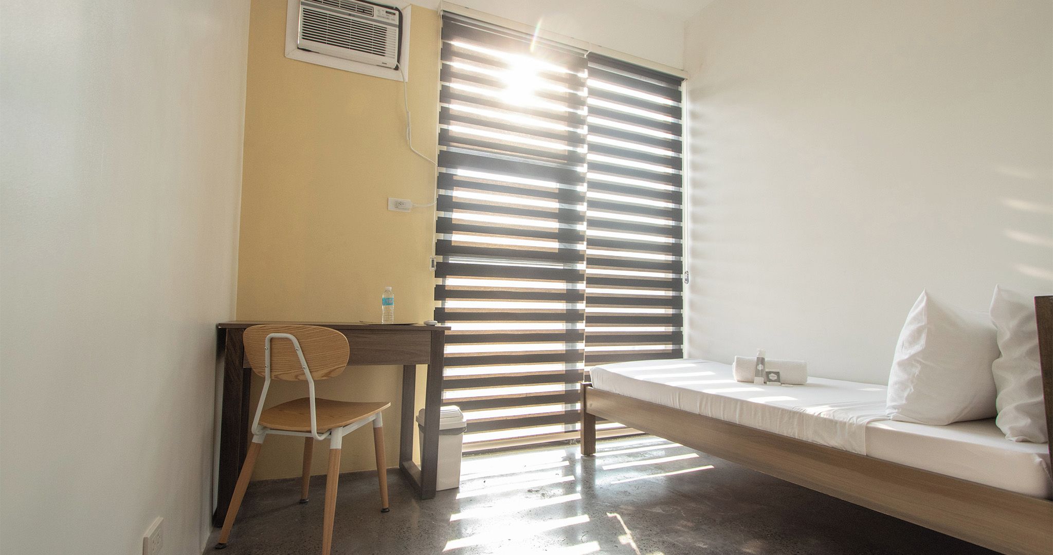 Ein kleines Zimmer mit Bett, Schreibtisch und Stuhl. Es gibt ein Fenster mit Jalousien, eine Wand-Klimaanlage und eine Wasserflasche auf dem Schreibtisch.